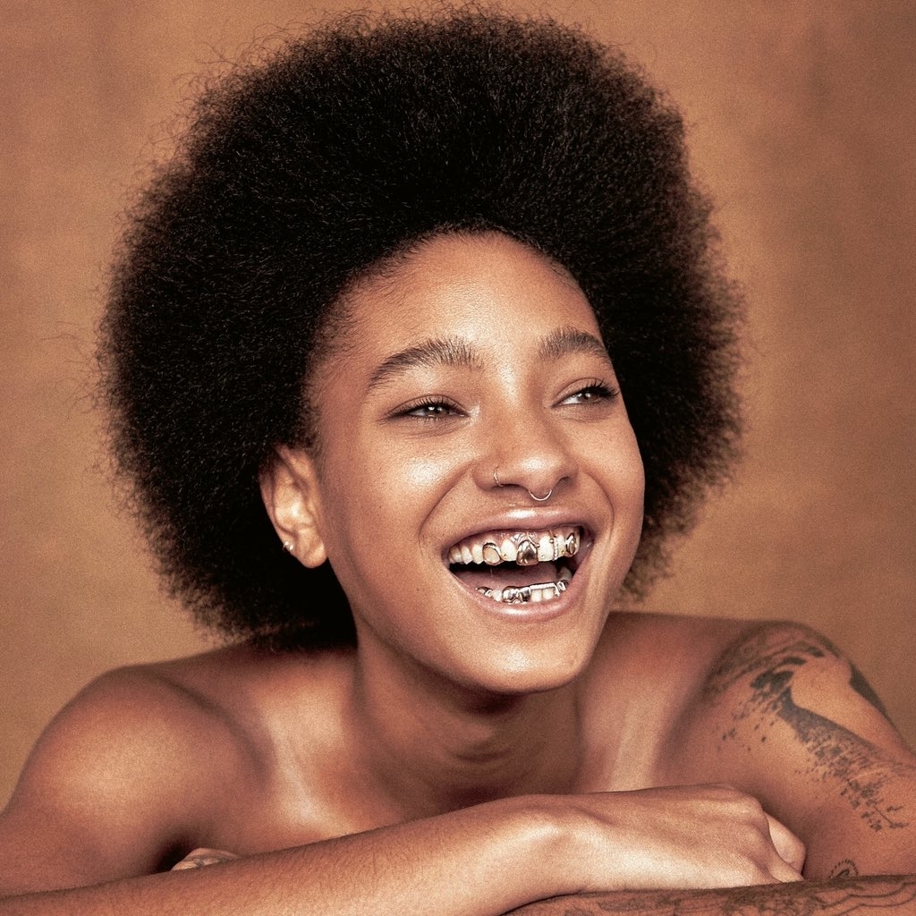 Willow smiles widely with an afro hairstyle, showing their teeth adorned with unique jewelry. She has tattoos on their shoulder and is resting her arms on a surface