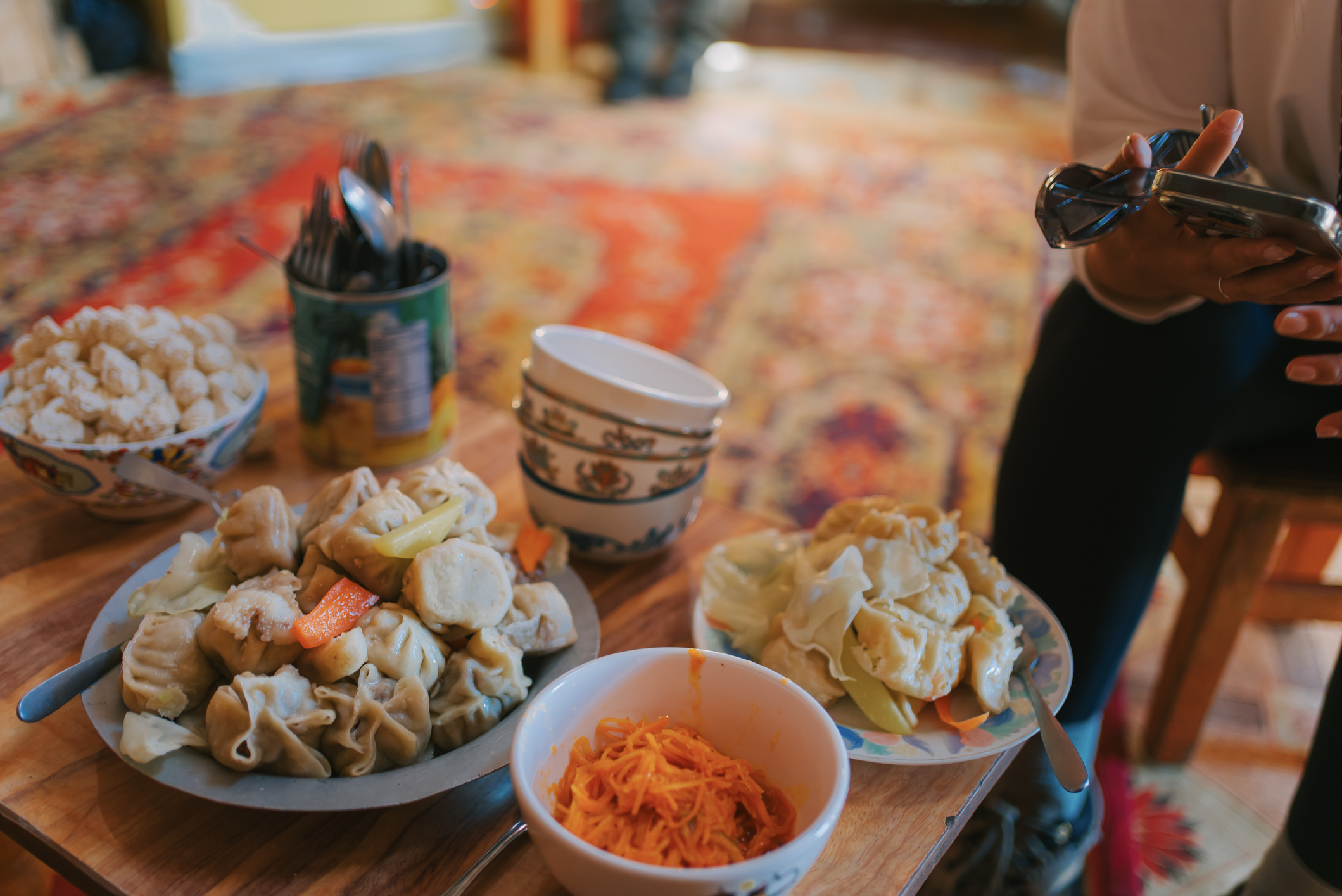 Various dumplings, pickled vegetables, and other dishes on a wooden table. A person holding a phone is partially visible on the right