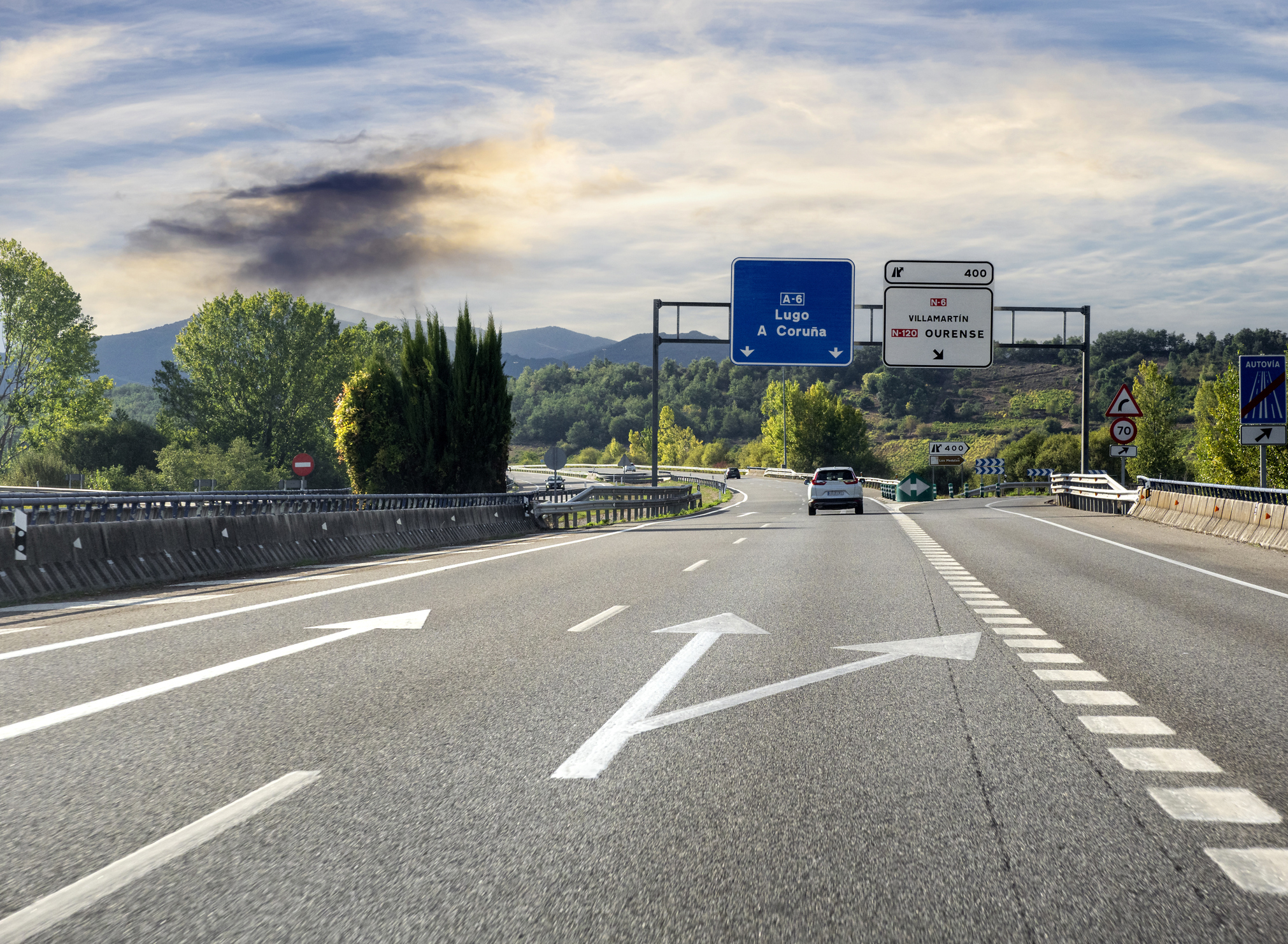 A highway in a scenic, hilly area with road signs directing to Lugo and A Coruna. A car travels on the lane with large white arrows painted on the road