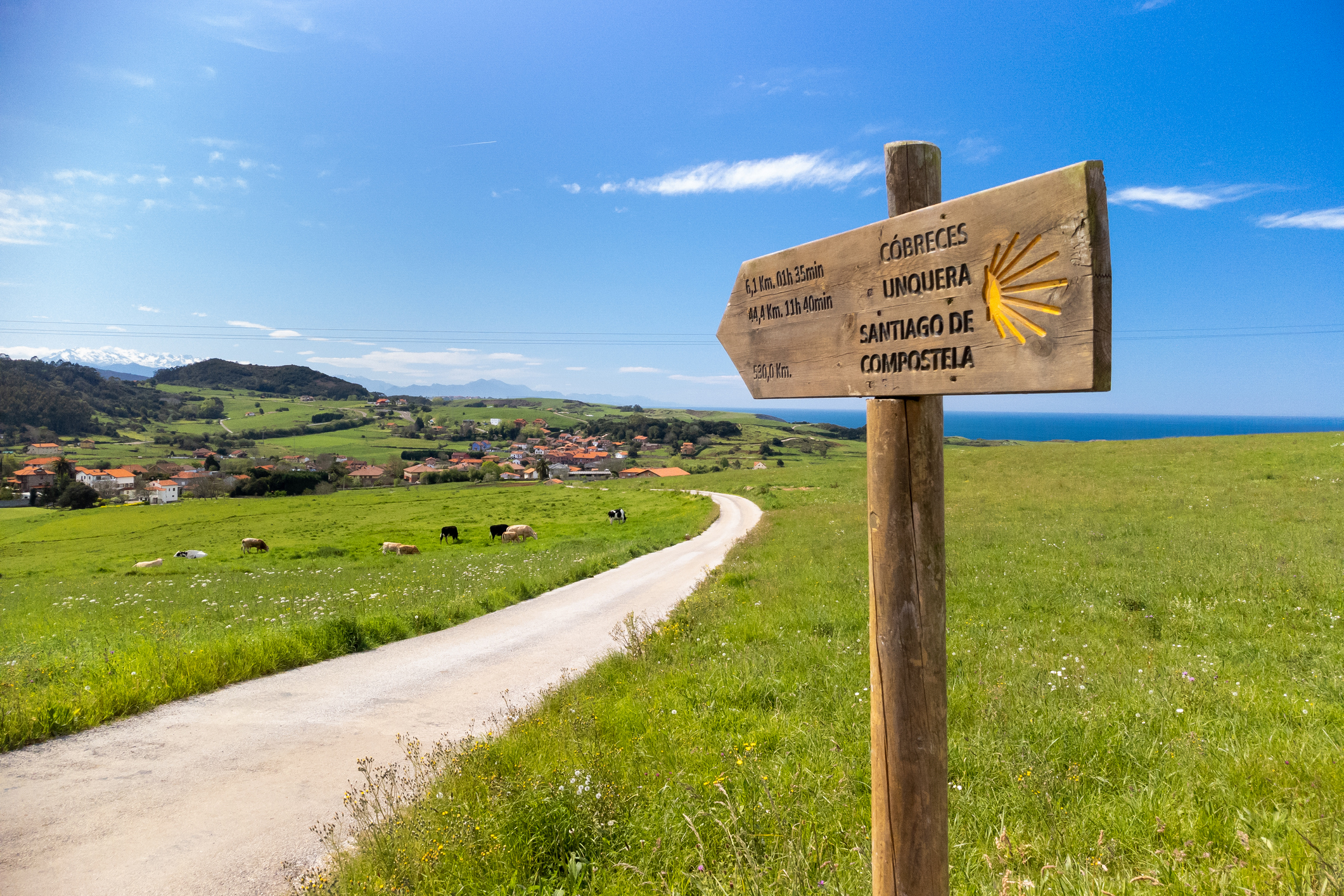 A rural road stretches through green fields towards a distant village, with a wooden signpost indicating directions to various destinations, including Santiago de Compostela