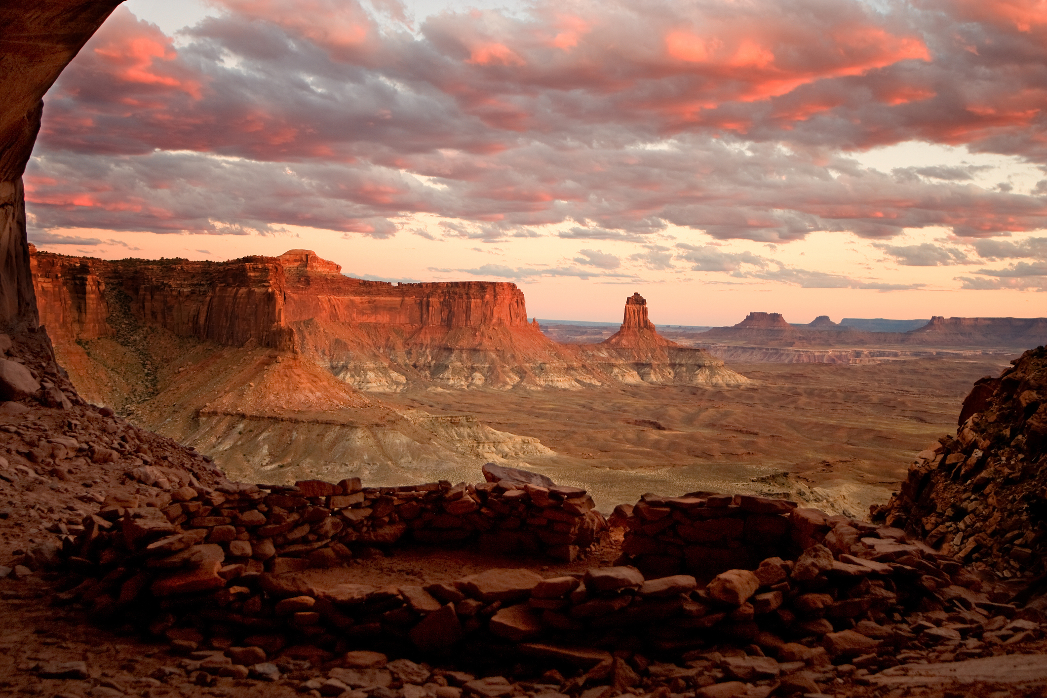 A breathtaking landscape featuring a view of rocky mesas and plateaus at sunrise or sunset, seen from a cave or sheltered space in the foreground