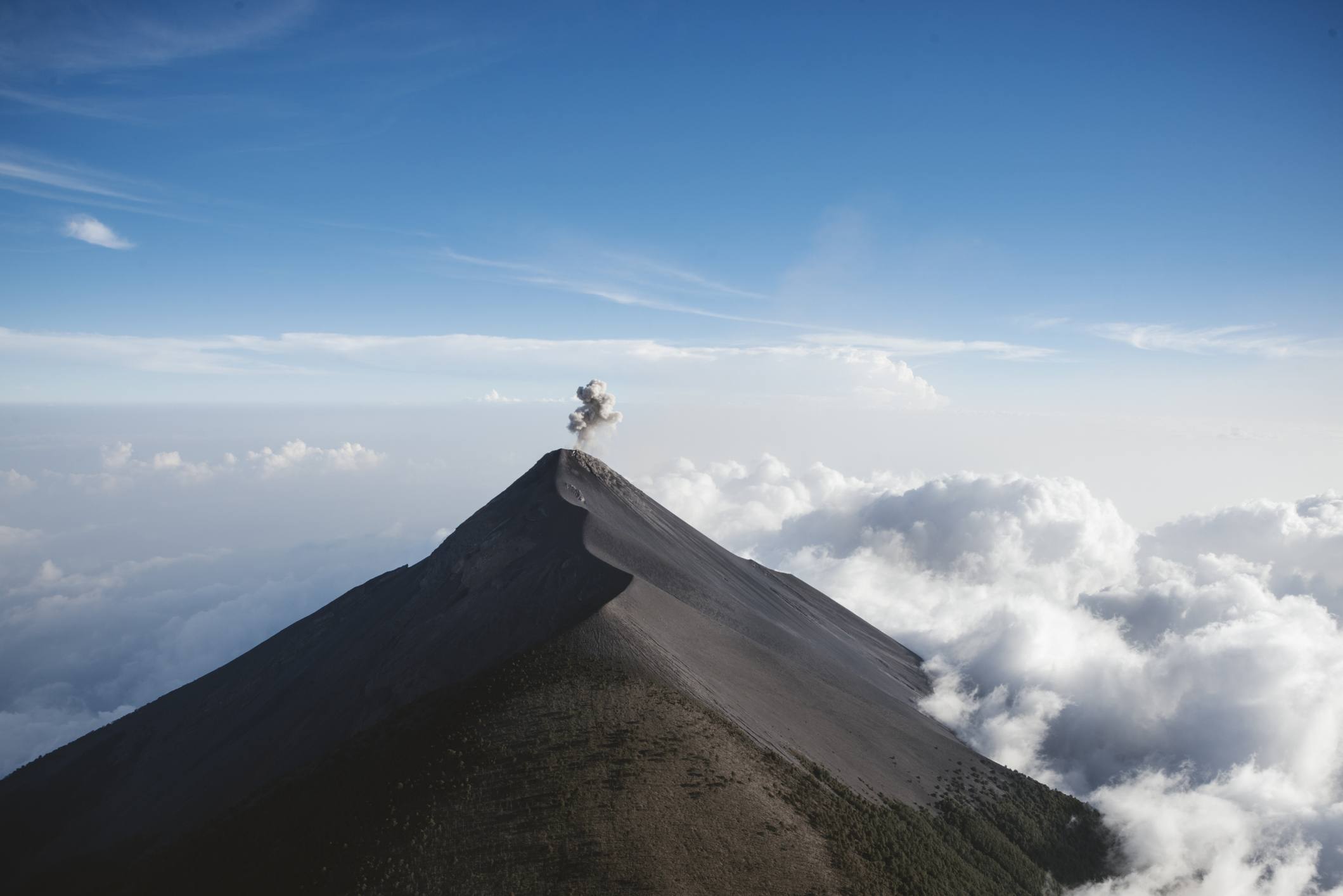 A volcano releasing smoke from its peak, surrounded by clouds and a clear sky above