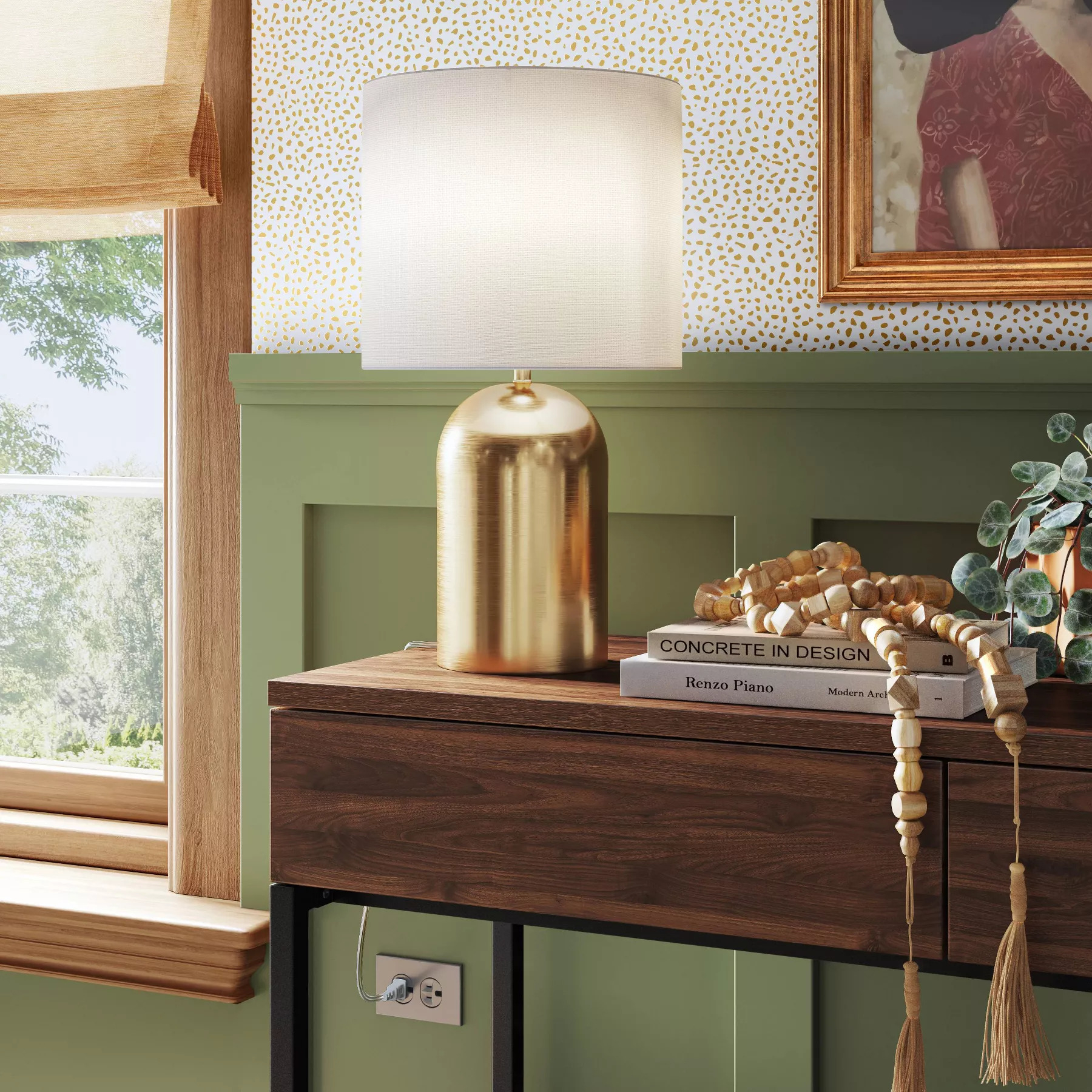 Lamp with white shade and gold body on wooden console table, alongside stacked books, decorative beads, and a plant