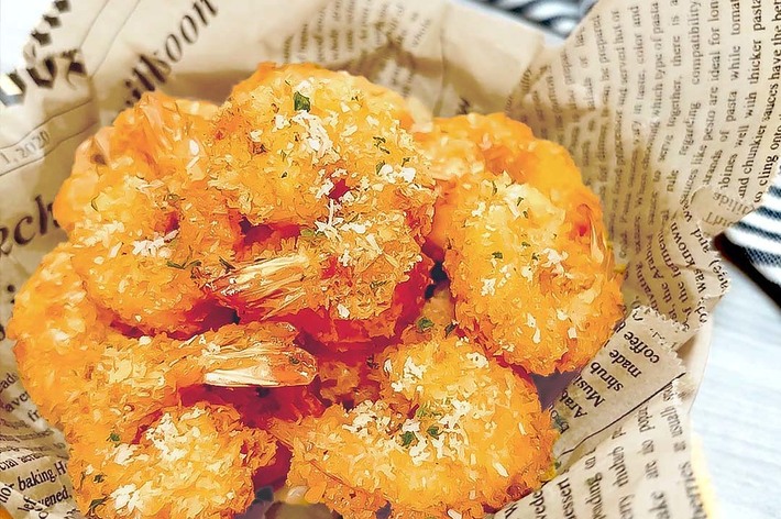 バットの上に並べられたパン粉で揚げられたエビフライ。背景には新聞紙風の包装が見える。