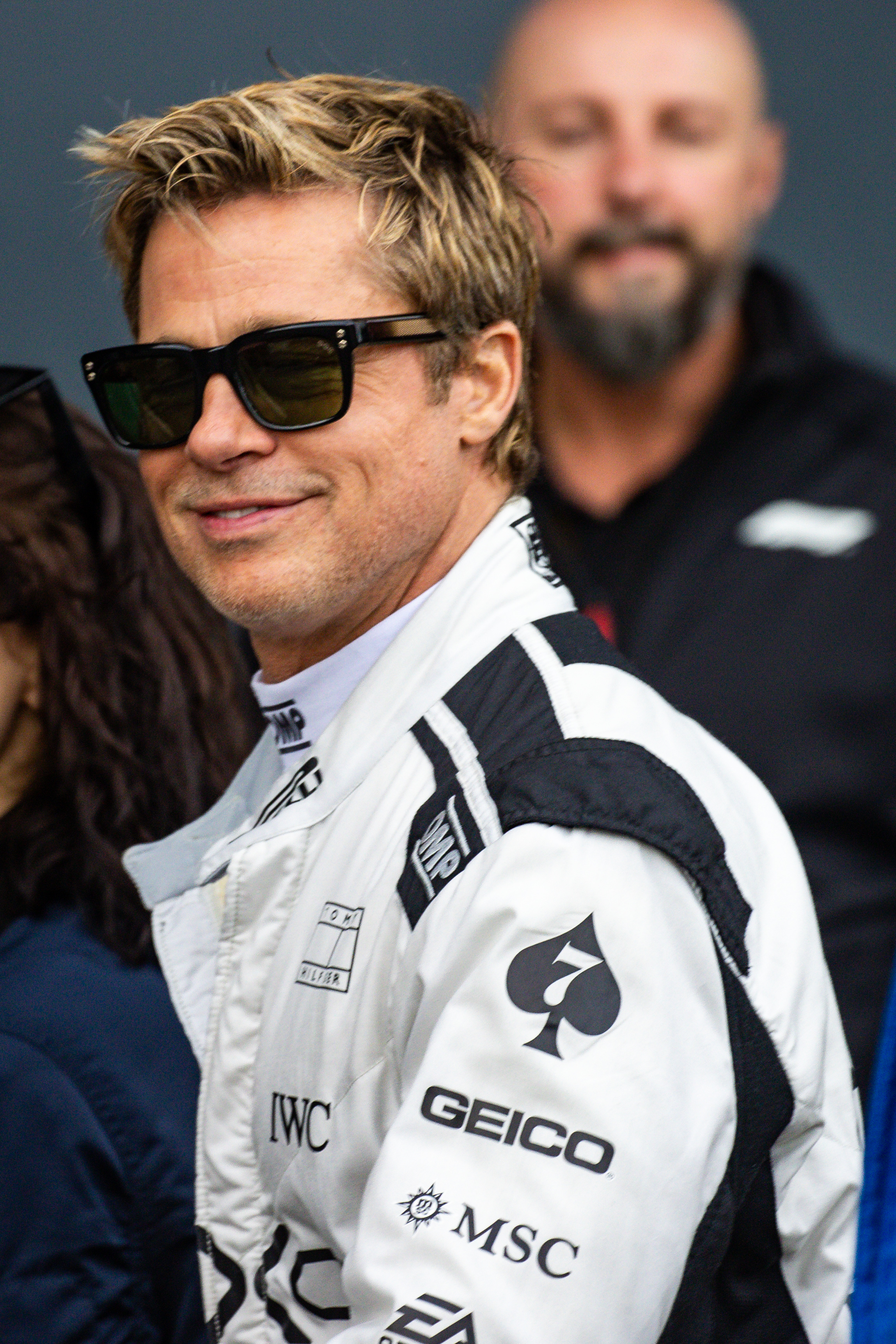 Brad Pitt wearing a racer jumpsuit with branded patches, and sunglasses, smiles while standing in a group