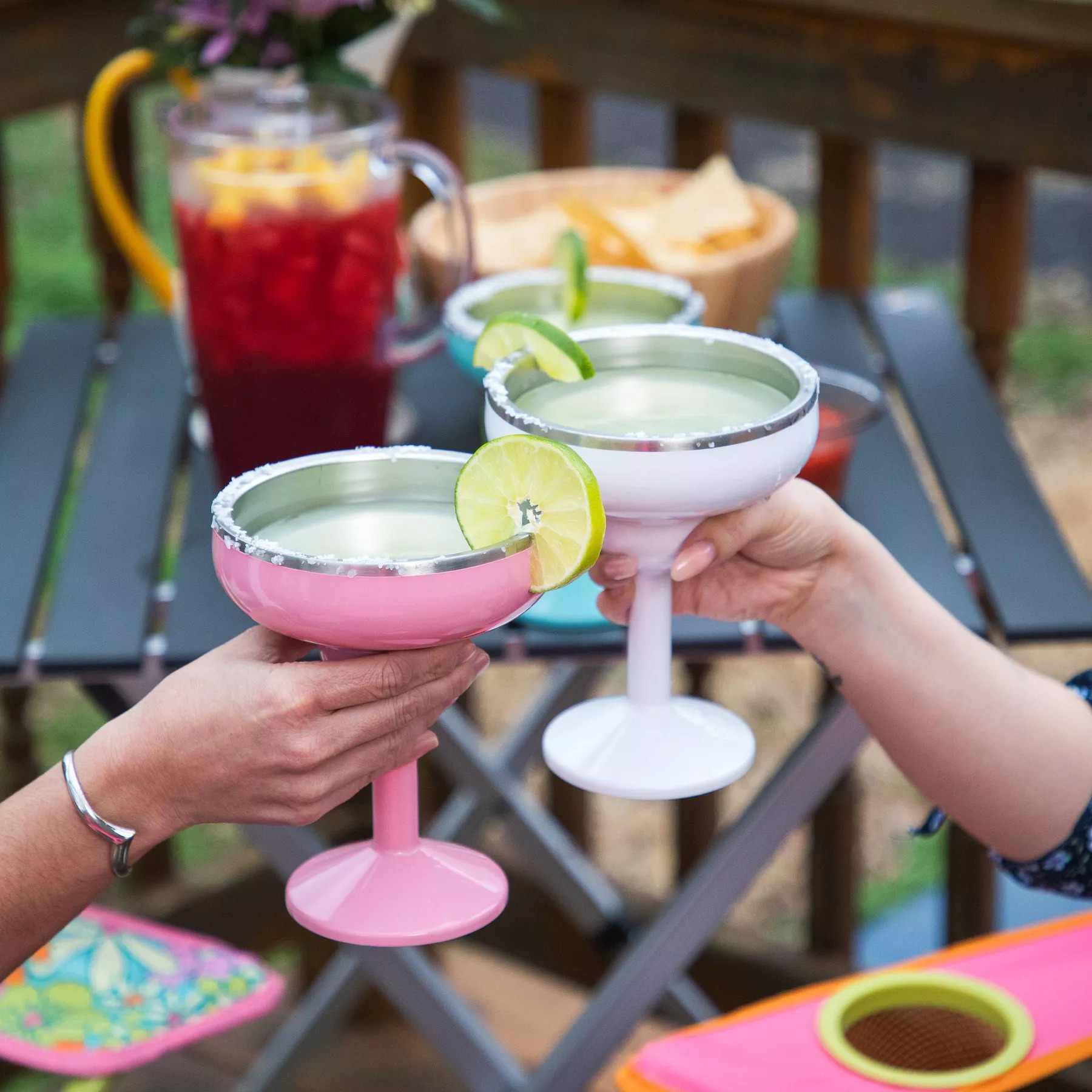 Two hands hold up pink and white margarita tumblers with lime wedges in a celebratory toast.