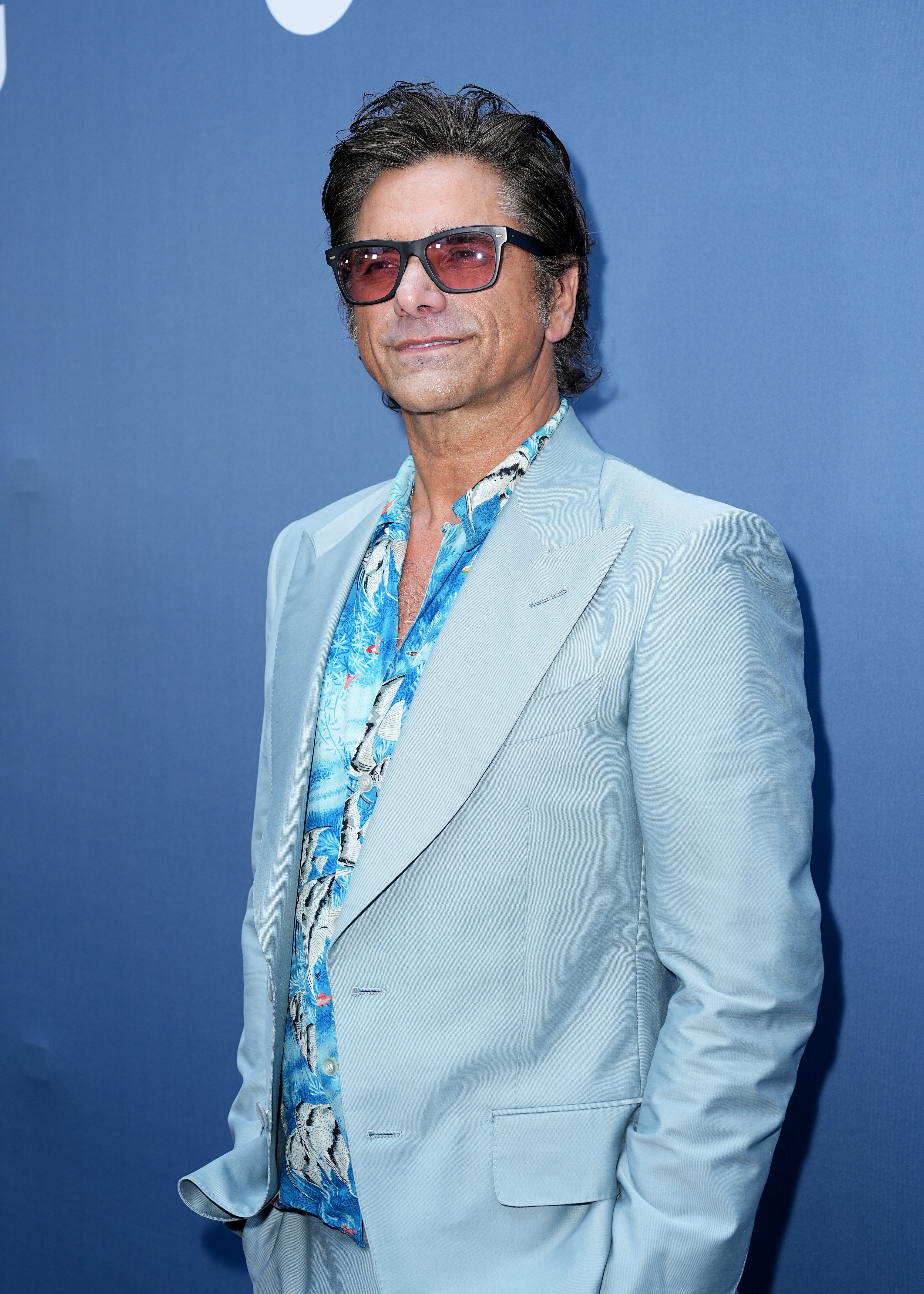 John Stamos wearing a light blue suit paired with a patterned shirt and sunglasses