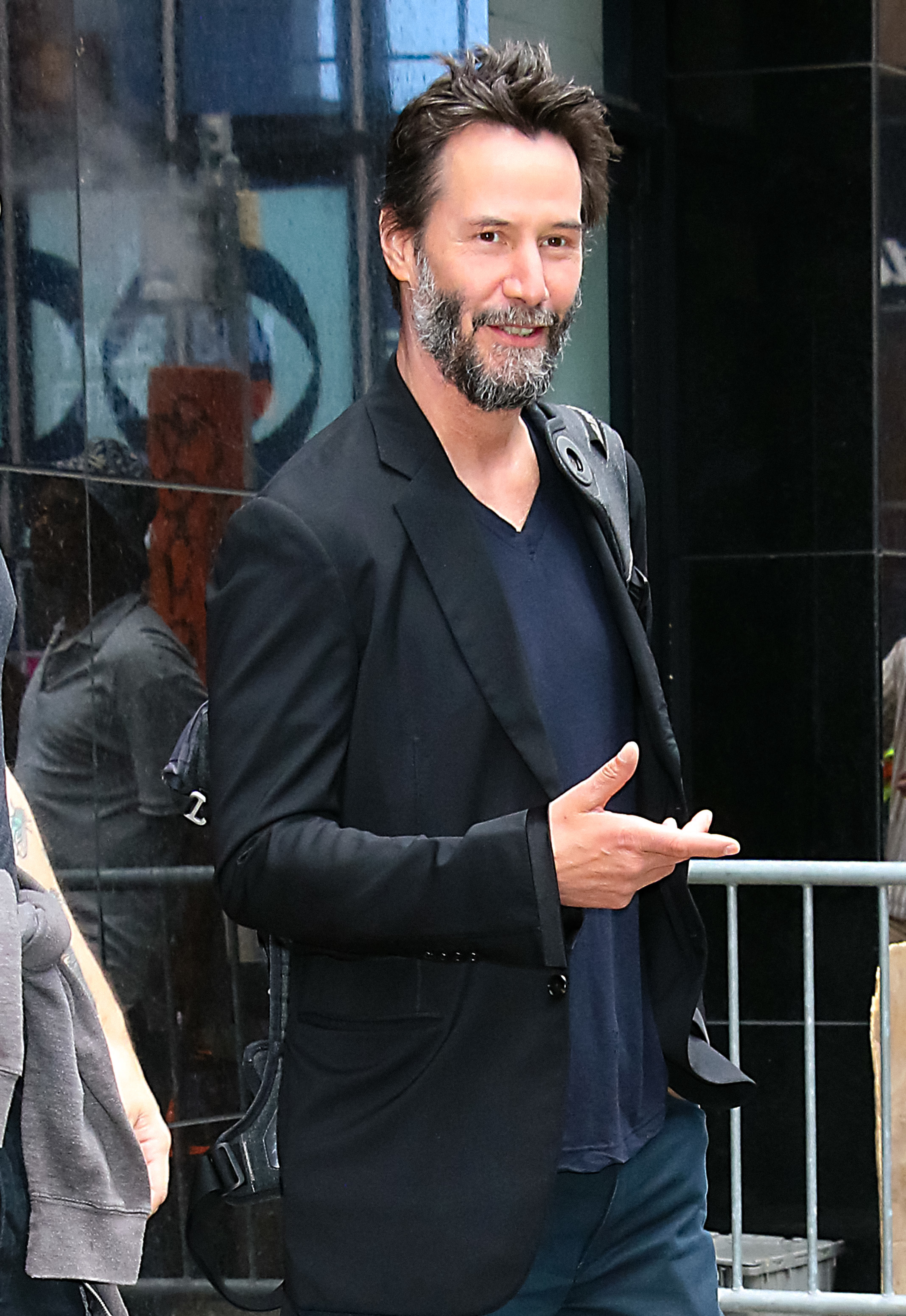 Keanu Reeves posing outdoors in a casual black blazer over a dark shirt, with a light smile, pointing with his right hand