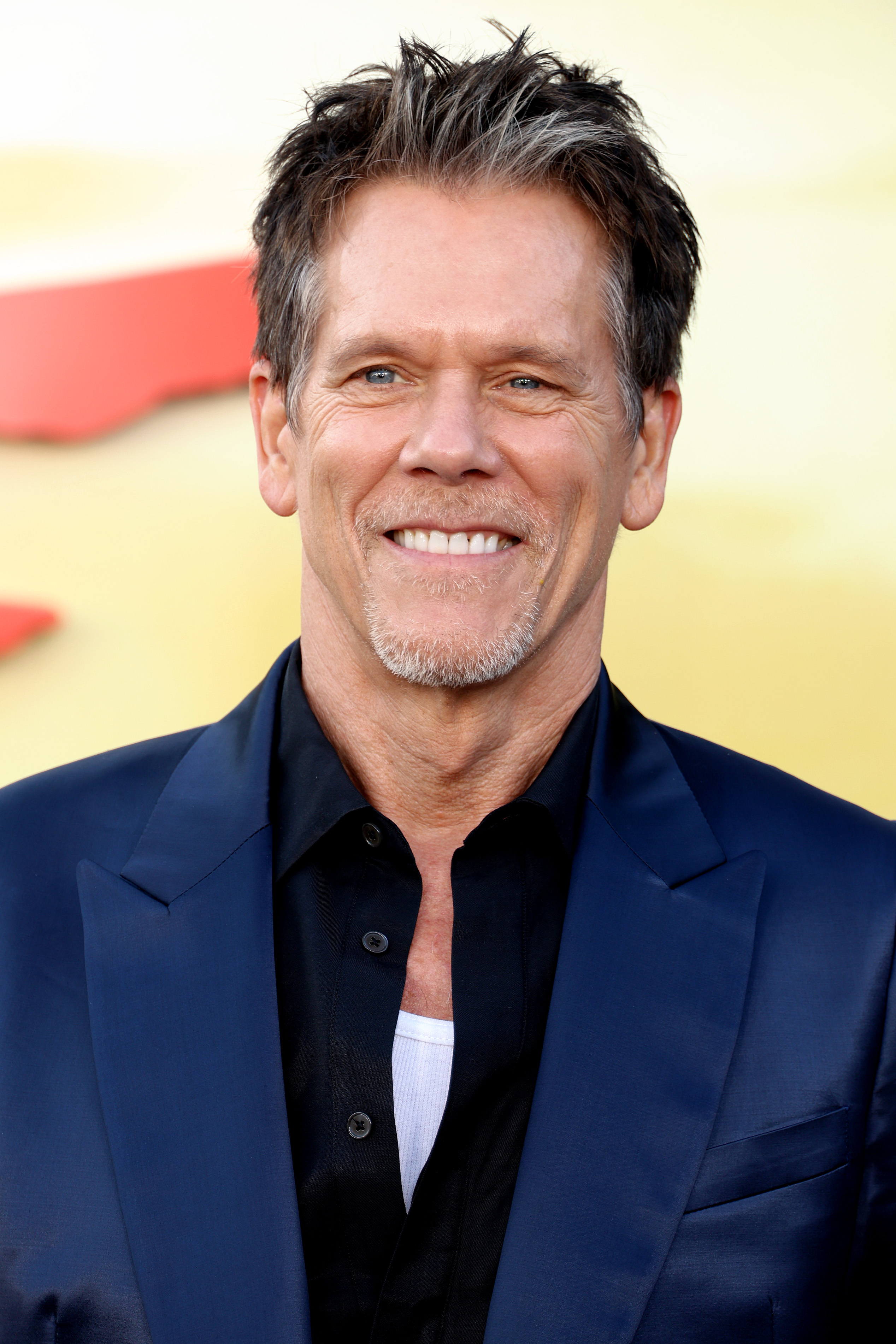 Kevin Bacon smiles in a blue blazer over a black shirt with a white undershirt, attending a red carpet event