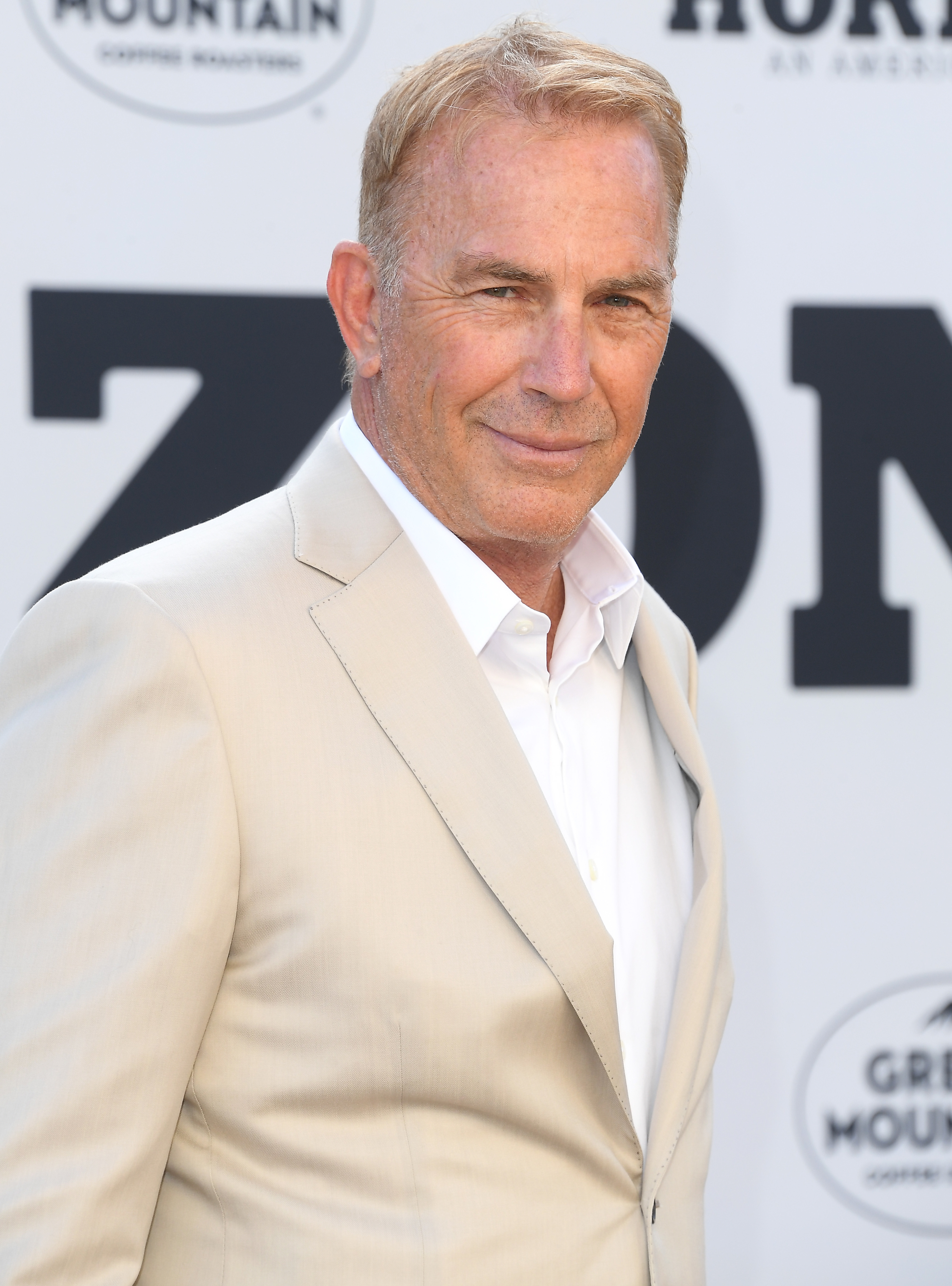 Kevin Costner in a beige suit with a white shirt, posing at an event