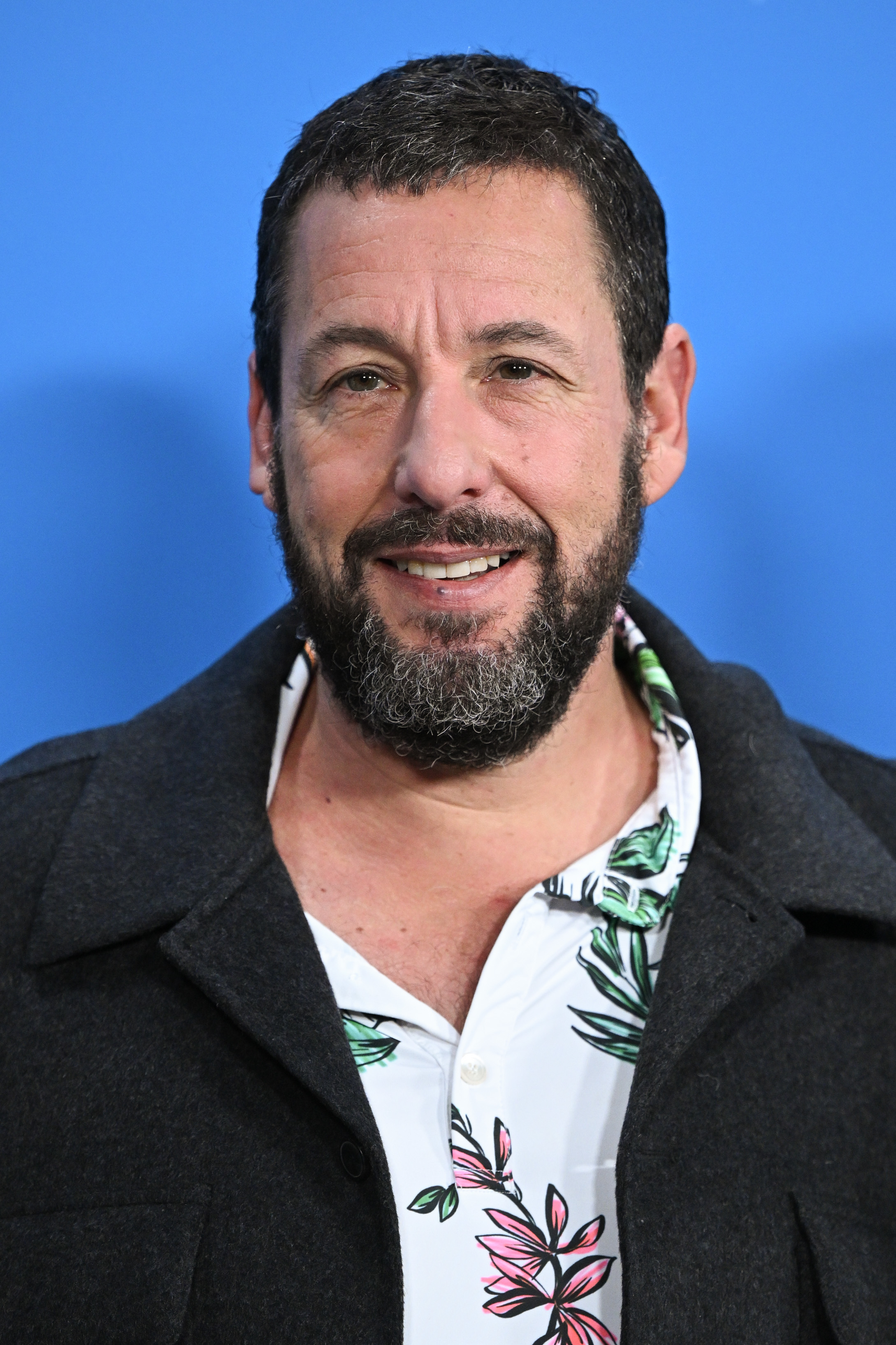 Adam Sandler smiling, wearing a casual coat over a floral shirt
