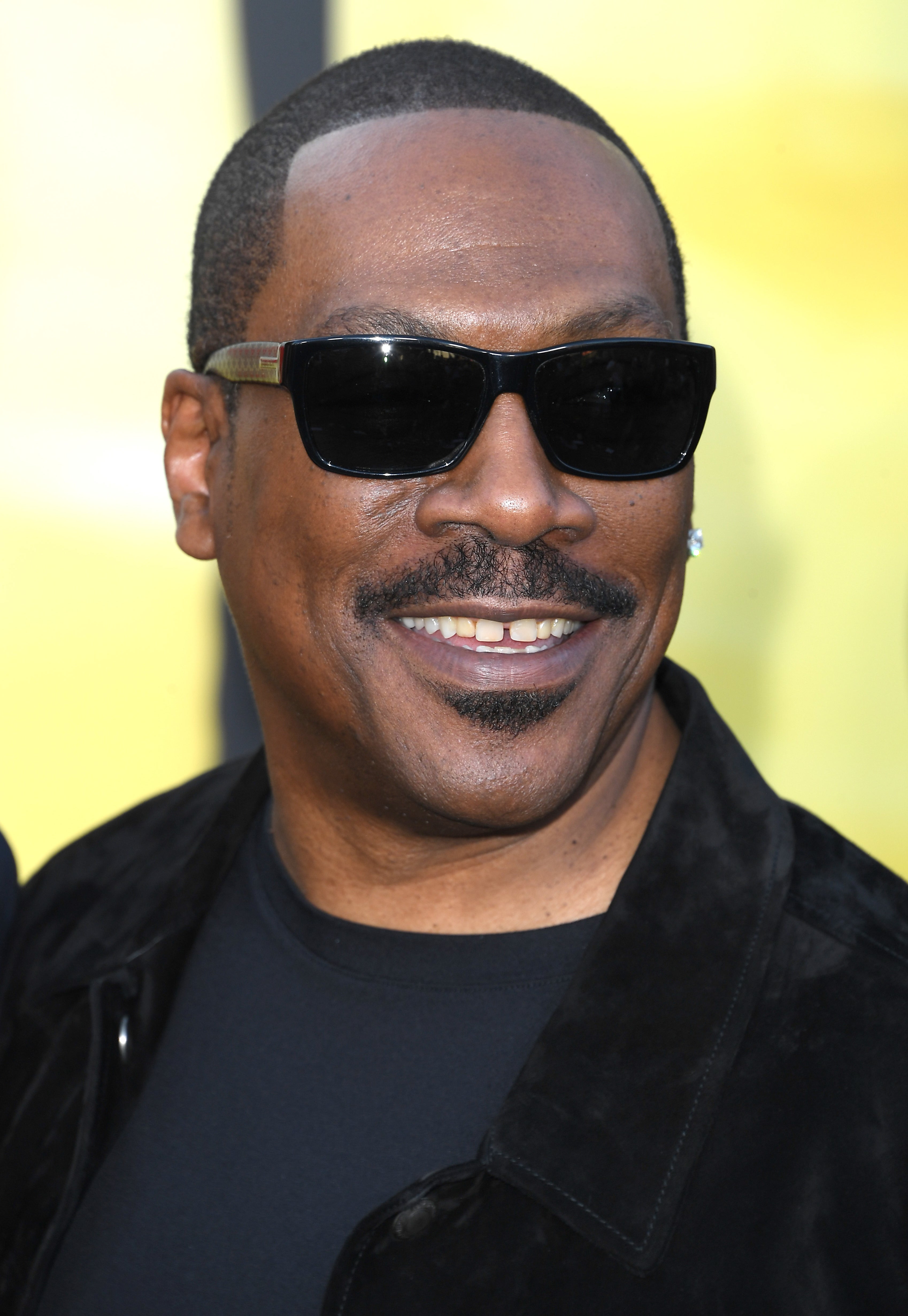 Eddie Murphy smiling and wearing black sunglasses, a black T-shirt, and a black jacket at a public event