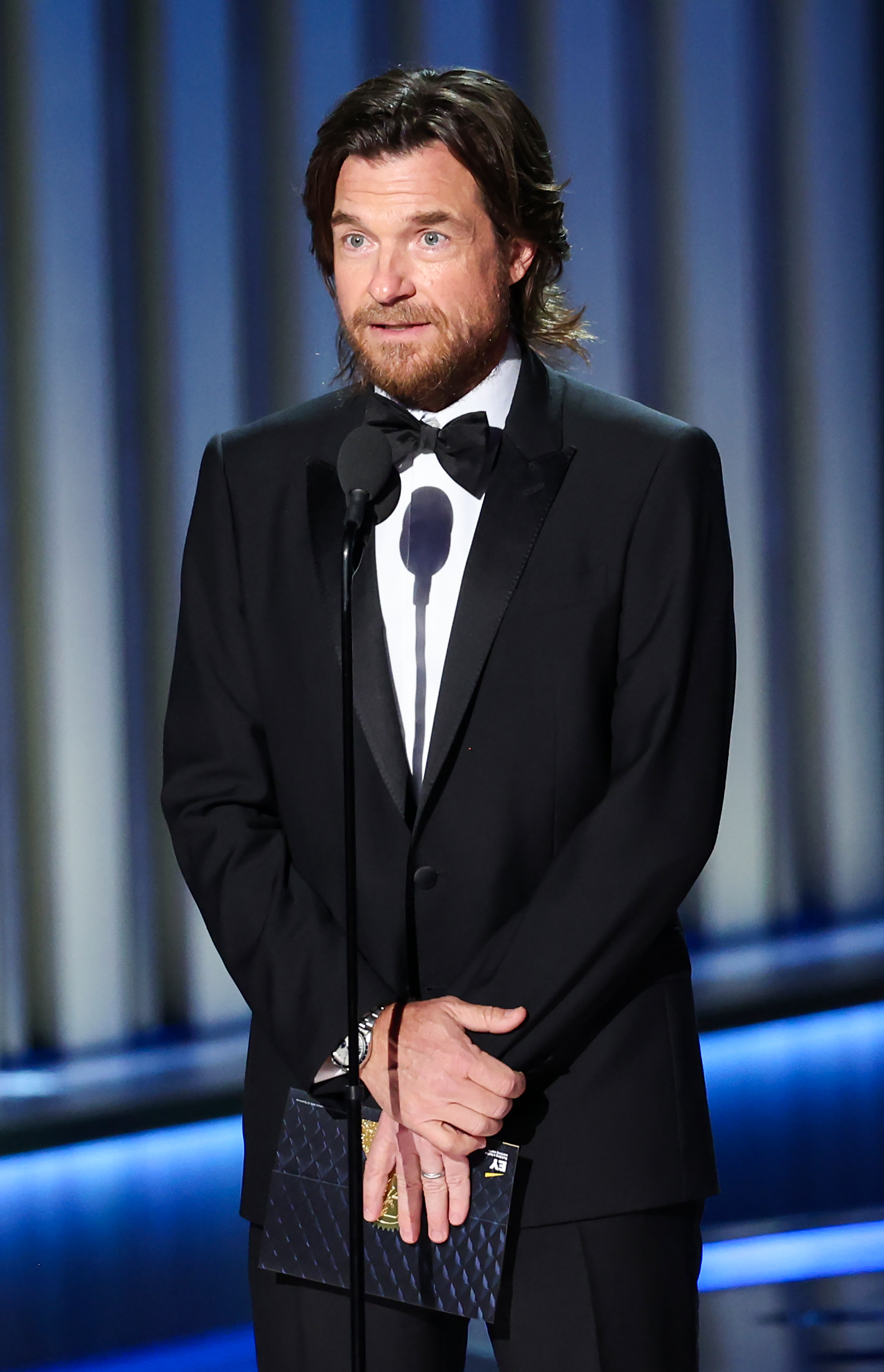 Jason Bateman in a black tuxedo with a bow tie, standing at a microphone on stage at an event