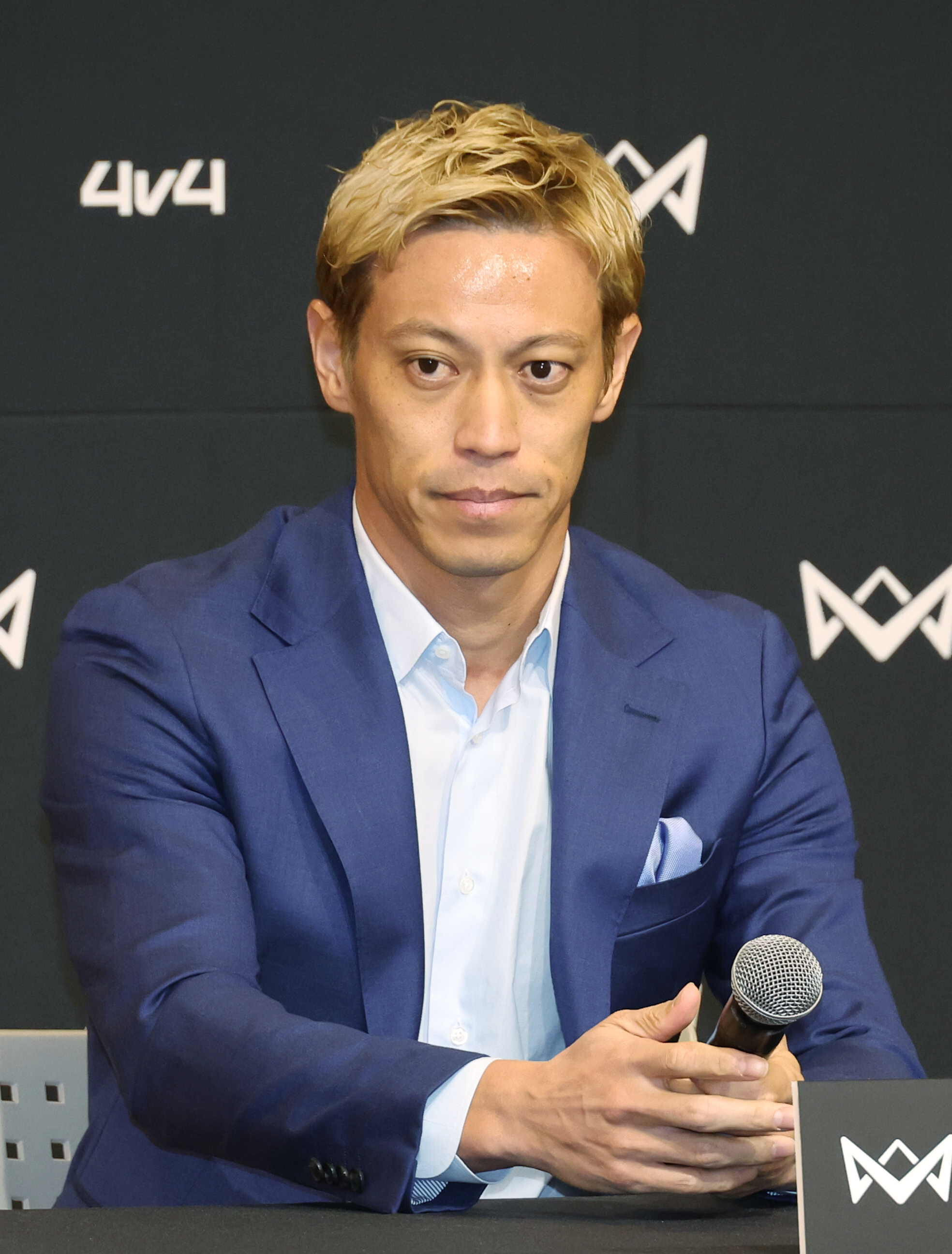 本田圭佑さんが記者会見でマイクを手にしている。青いスーツを着用。