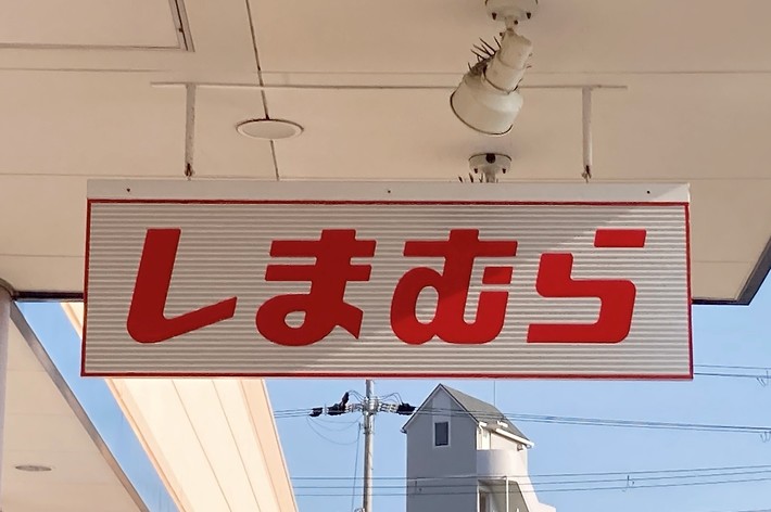 A store sign with Japanese characters reads: &ldquo;しまむら&rdquo;. The sky and part of a building are visible in the background