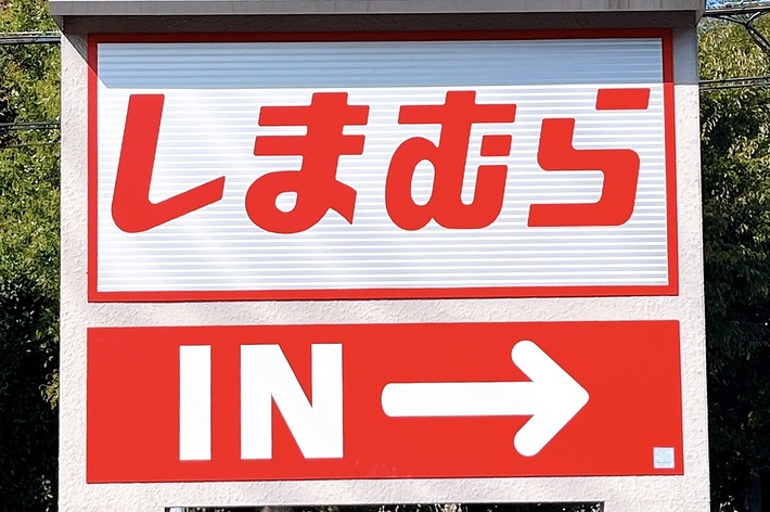 A sign in Japanese reads “Shimamura” with an arrow and “IN” indicating the entrance direction. A street and trees are in the background