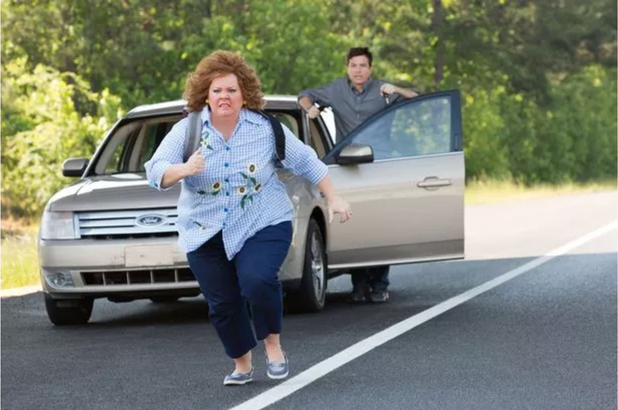 Melissa McCarthy runs away from a car while a man stands near the car with the driver-side door open on a road surrounded by trees