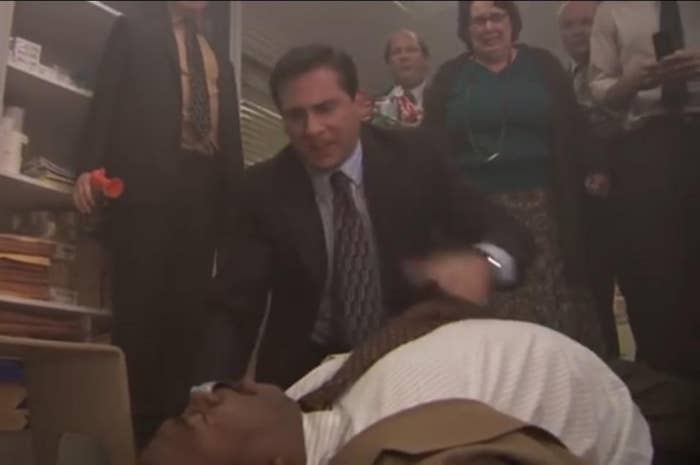 Steve Carell compresses a man's chest while onlookers, including Brian Baumgartner and Phyllis Smith, watch anxiously