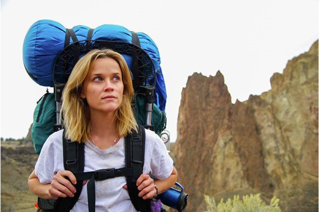 Reese Witherspoon hiking with a large backpack near a rocky terrain. She wears a simple t-shirt and looks thoughtful