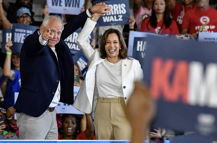 Tim Walz and Kamala Harris raising their arms and smiling at a rally, surrounded by supporters holding signs