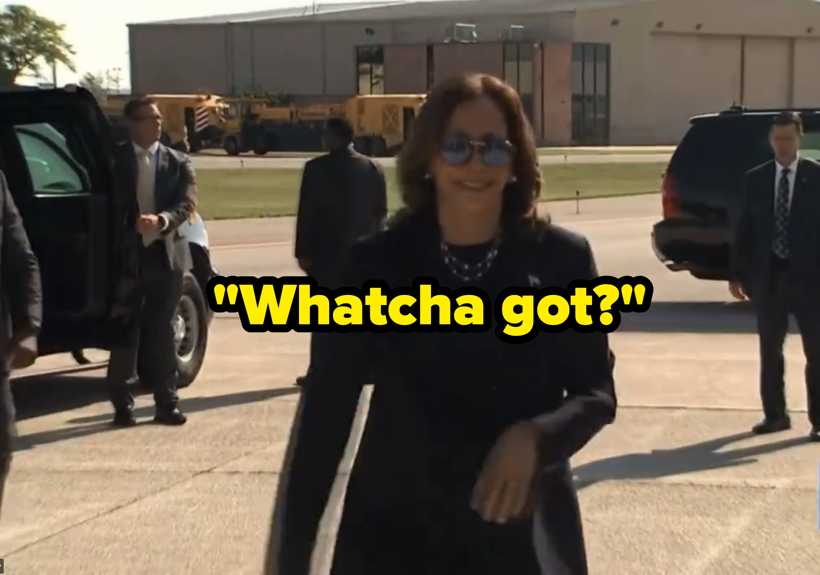 Kamala Harris, wearing a dark suit and sunglasses, smiles while walking on a tarmac with security personnel in the background