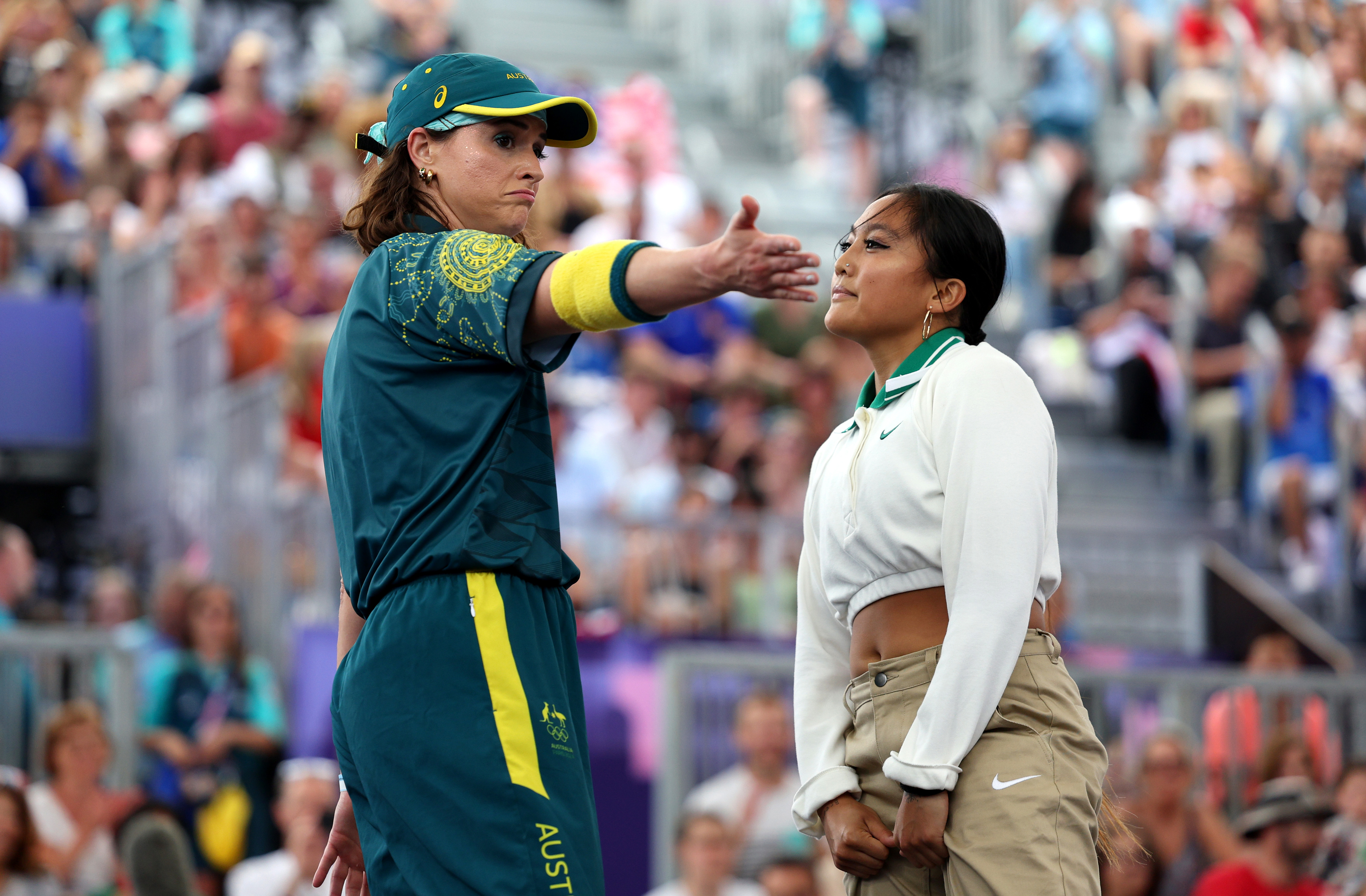 Beth Mooney in Australian cricket uniform extends her arm towards a person wearing a white top and beige pants on the cricket field, with a crowd in the background