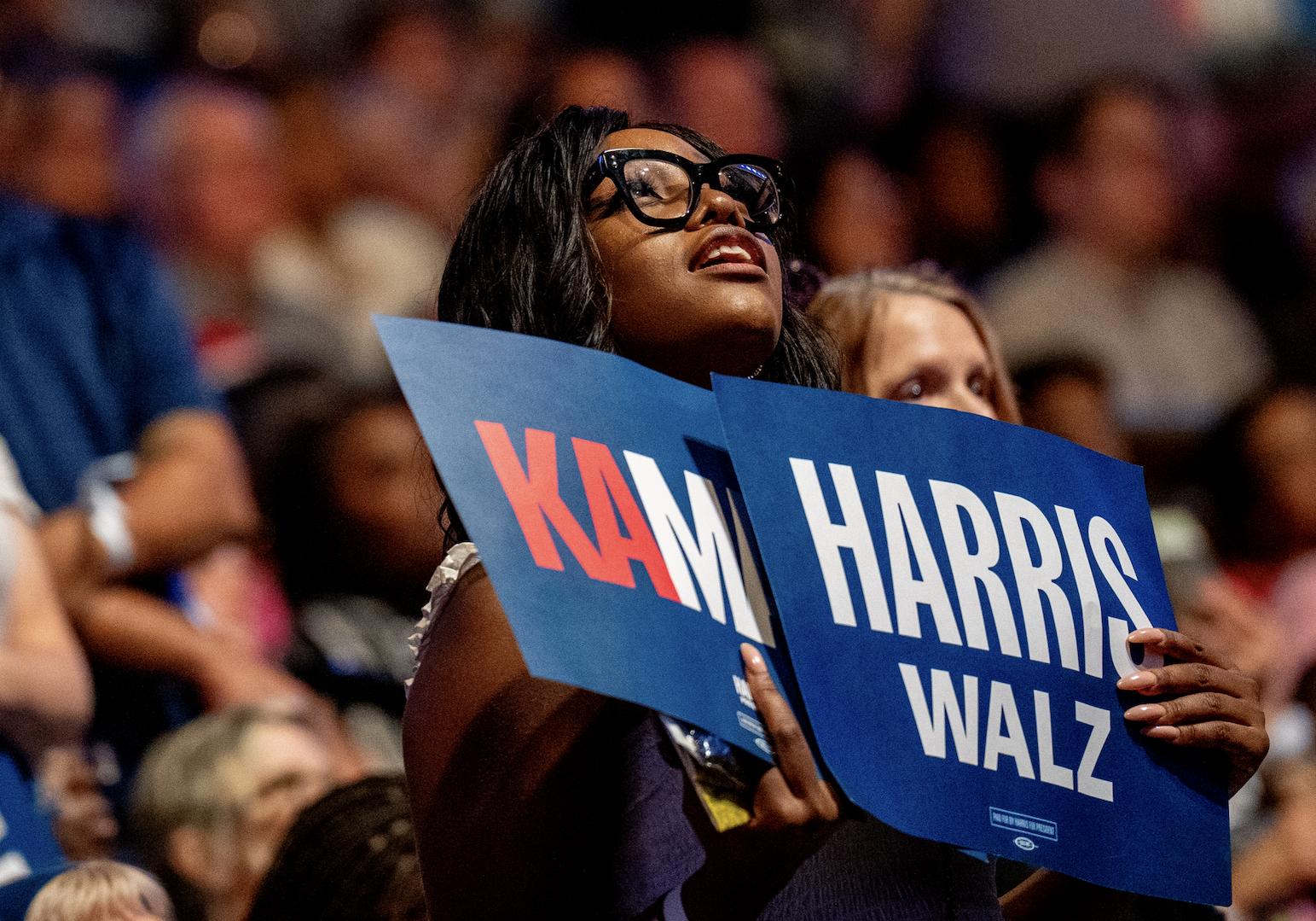 A person holds blue signs reading "KAM HARRIS WALZ" at a crowded event. The person is wearing large glasses and appears focused on the stage