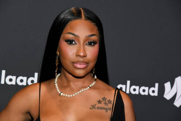 Yung Miami attends a GLAAD event, wearing a black dress with a pearl necklace and long, straight hair. She has star tattoos on her chest