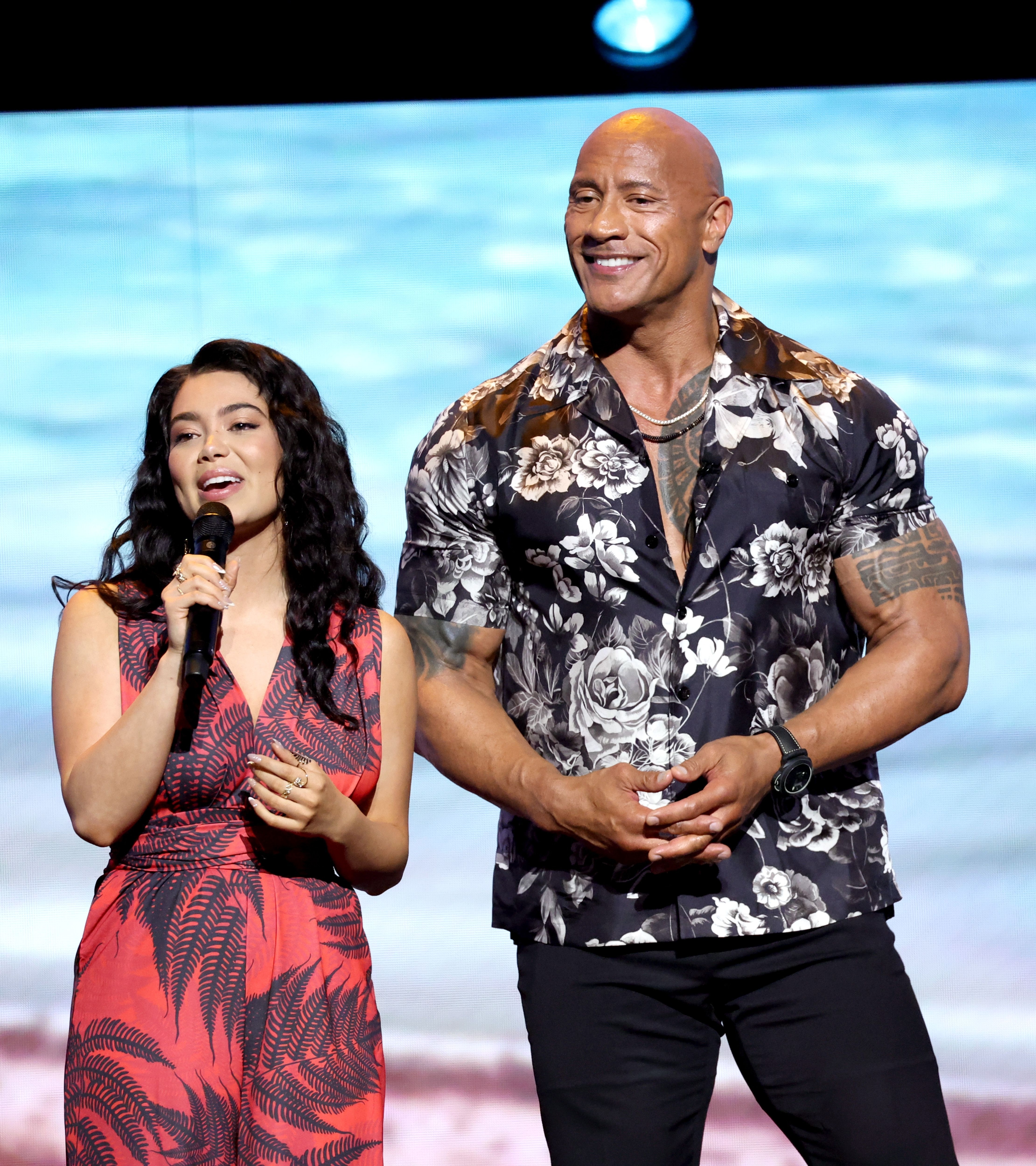 Auli'i Cravalho in a floral dress and Dwayne Johnson in a floral shirt speak on stage with a beach backdrop