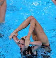 Five synchronized swimmers perform an intricate, acrobatic routine in a pool, with their legs raised above their heads in unison