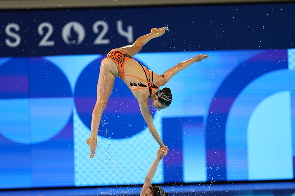 Synchronized swimmers, one lifted in the air by the other, perform during a competition. The background shows "2024". Names not known