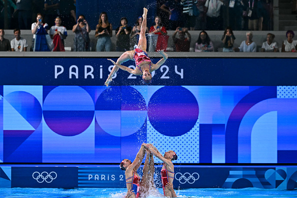 Synchronized swimming team performs an acrobatic routine at the Paris 2024 Olympics, with one swimmer being launched into the air. Audience watches in the background