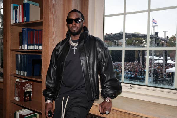 Sean &quot;Diddy&quot; Combs leans against a library window in a black leather jacket, dark sunglasses, and jewelry, with a crowd and stage visible outside