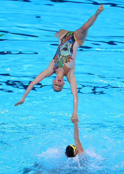Synchronized swimmers perform an acrobatic lift in a pool. One swimmer is held upside down in mid-air by another's hand. Names of persons are not known
