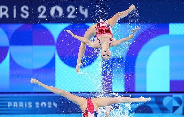 Two synchronized swimmers wearing red outfits perform a mid-air flip during a routine at the Paris 2024 event