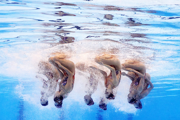 Synchronized swimming team performs a routine underwater, legs extended vertically with water splashes, each swimmer in alignment. Names of the team members are not provided