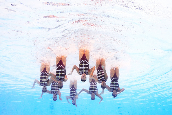 Synchronized swimmers perform an underwater routine, clad in matching one-piece swimsuits with geometric patterns