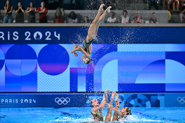 Athletes perform an acrobatic routine in the pool during the Paris 2024 Olympics, with one gymnast airborne above the others who support her