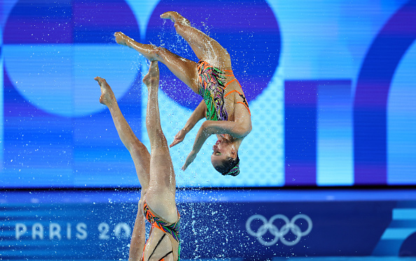 Two synchronized swimmers perform a choreographed routine at the Paris 2024 Olympics, mid-air with water splashing around them