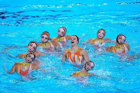 Eight synchronized swimmers perform an artistic routine in a pool, wearing matching swimsuits and makeup