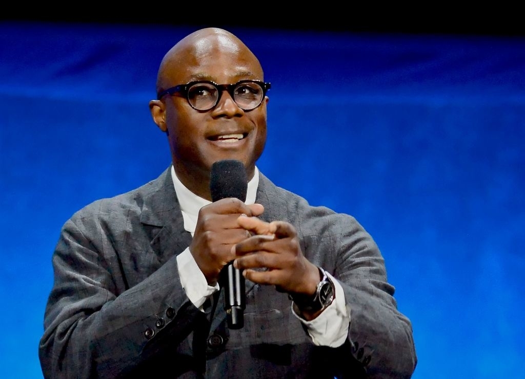 Man wearing glasses and a suit speaks into a microphone while gesturing with his other hand against a blue background