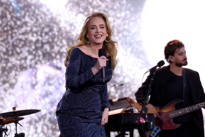 Adele sings on stage in an elegant, patterned dress, holding a microphone. A guitarist plays in the background