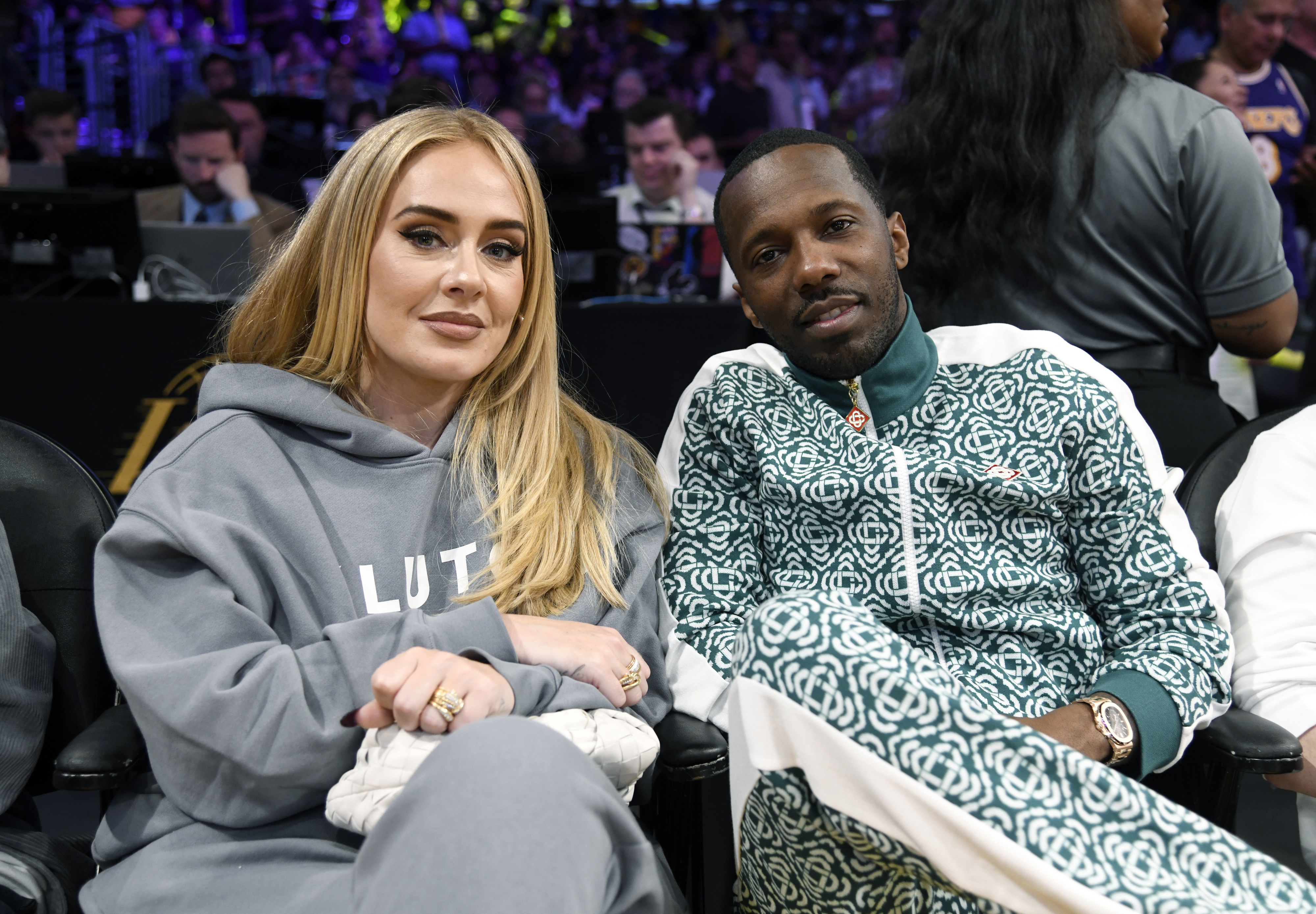 Adele and Rich Paul are sitting together at a basketball game. Adele is wearing a hoodie, and Rich Paul is wearing a patterned tracksuit