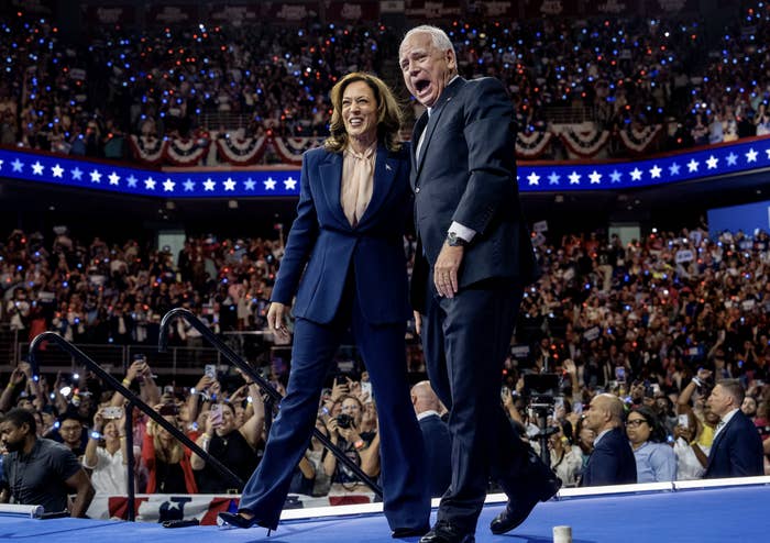 Kamala Harris and Doug Emhoff at a large, indoor rally, smiling and waving to the crowd from a stage adorned with patriotic decorations