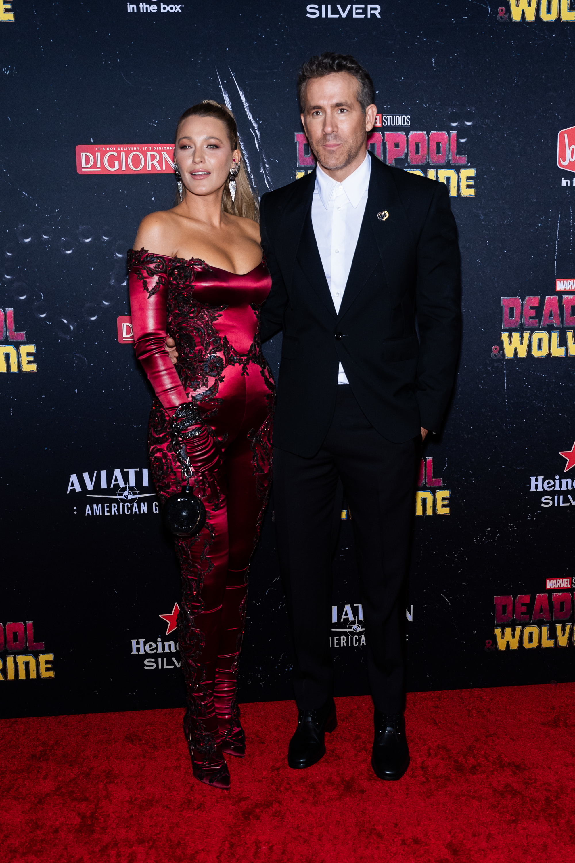 Blake Lively, in a form-fitting, off-shoulder red jumpsuit, and Ryan Reynolds, in a black suit with a white shirt, pose on the Deadpool 3 red carpet