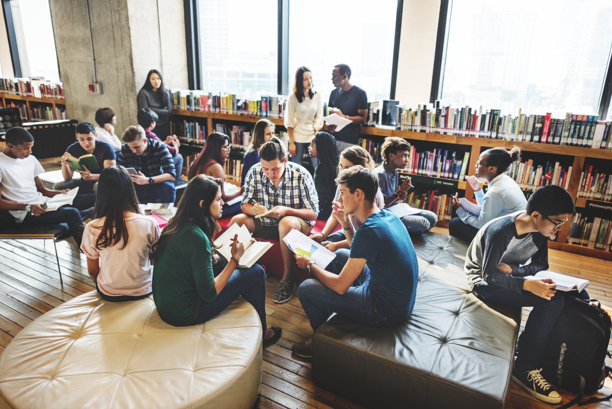 People seated in groups reading and discussing in a library with bookshelves and large windows in the background