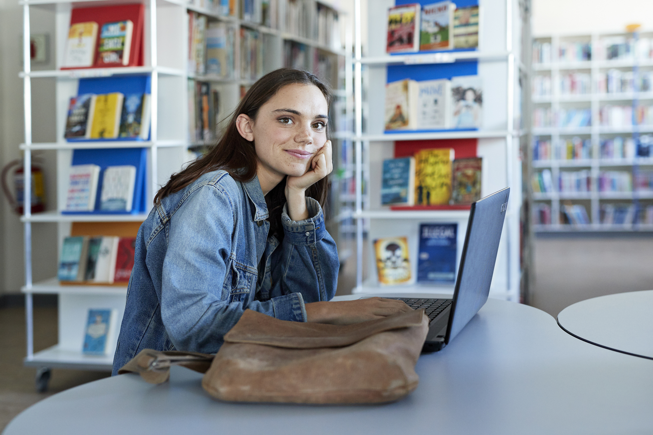 A woman in a denim jacket is seated at a laptop in a library. She is looking at the camera, with her chin rested on her hand, next to a brown bag