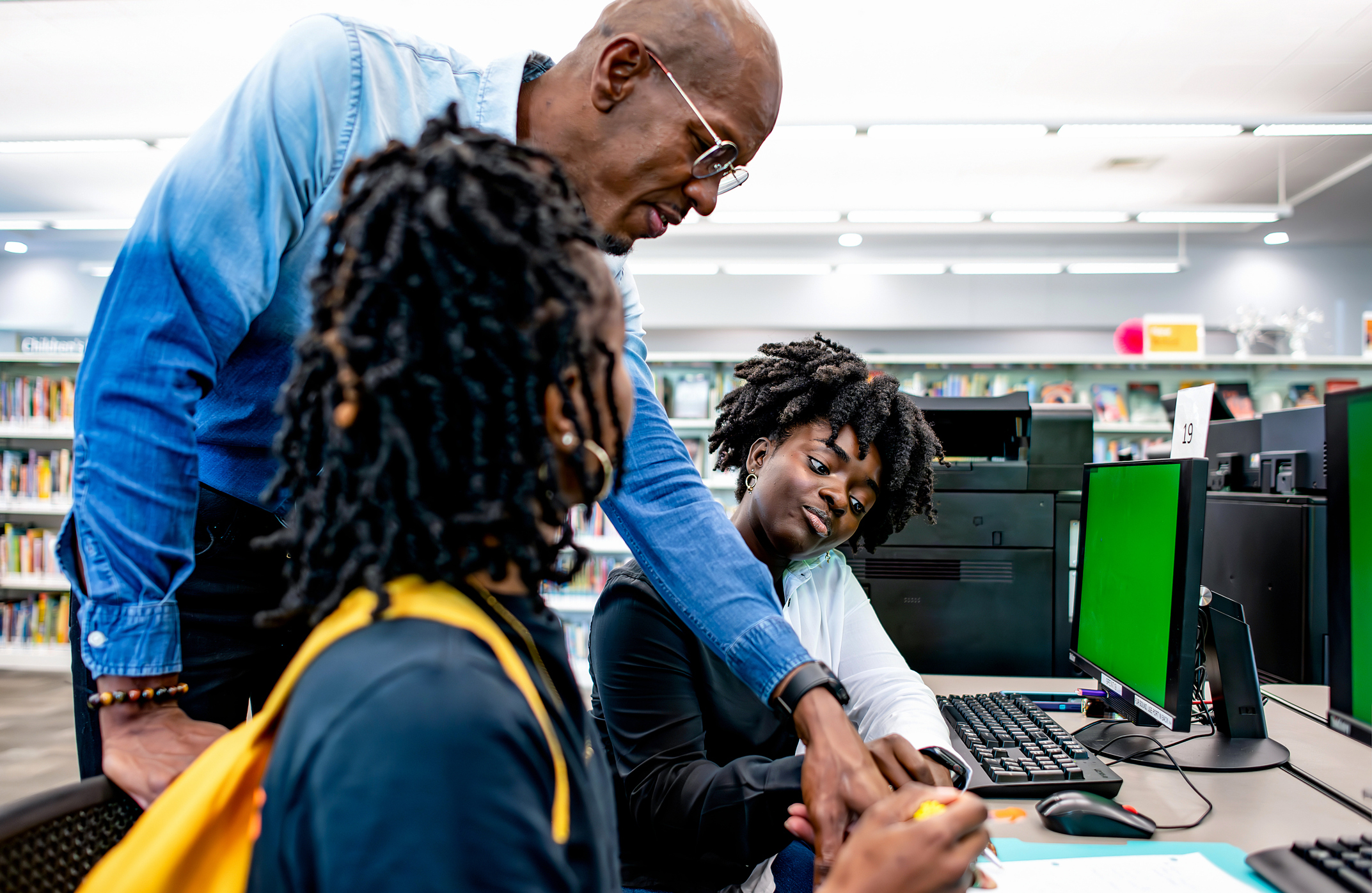 An instructor assists two students with their work on computers in a library setting