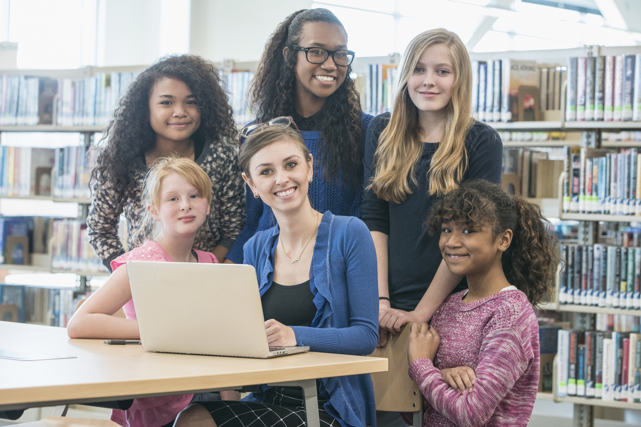 A woman sits at a desk with a laptop, surrounded by five young girls, all smiling, in a library setting with shelves of books in the background