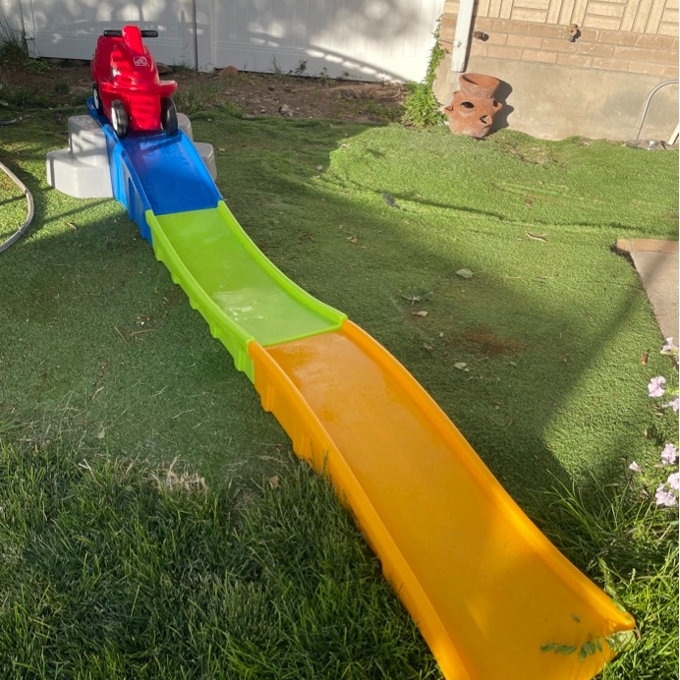 A red toy car sits at the top of a colorful plastic slide in a backyard, ready to roll down the blue, green, and yellow track laid out on the grass